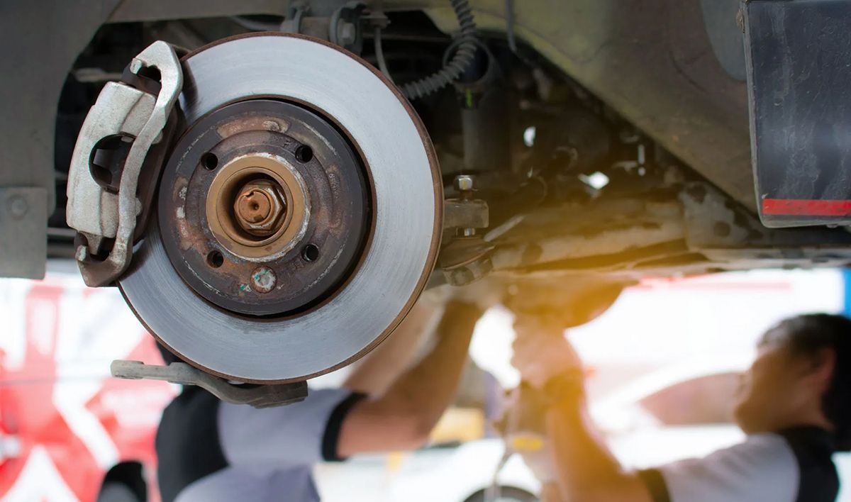 Car brake disc and caliper; mechanics working underneath, blurred background.