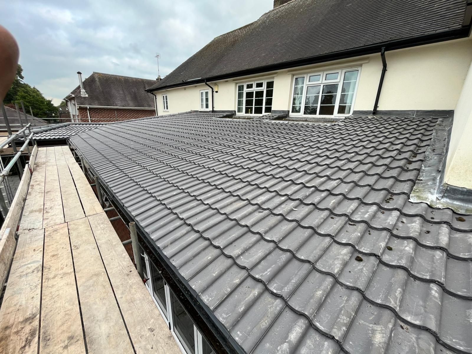 Gray tiled roof of a building; scaffolding at left, windows in background, cloudy sky.