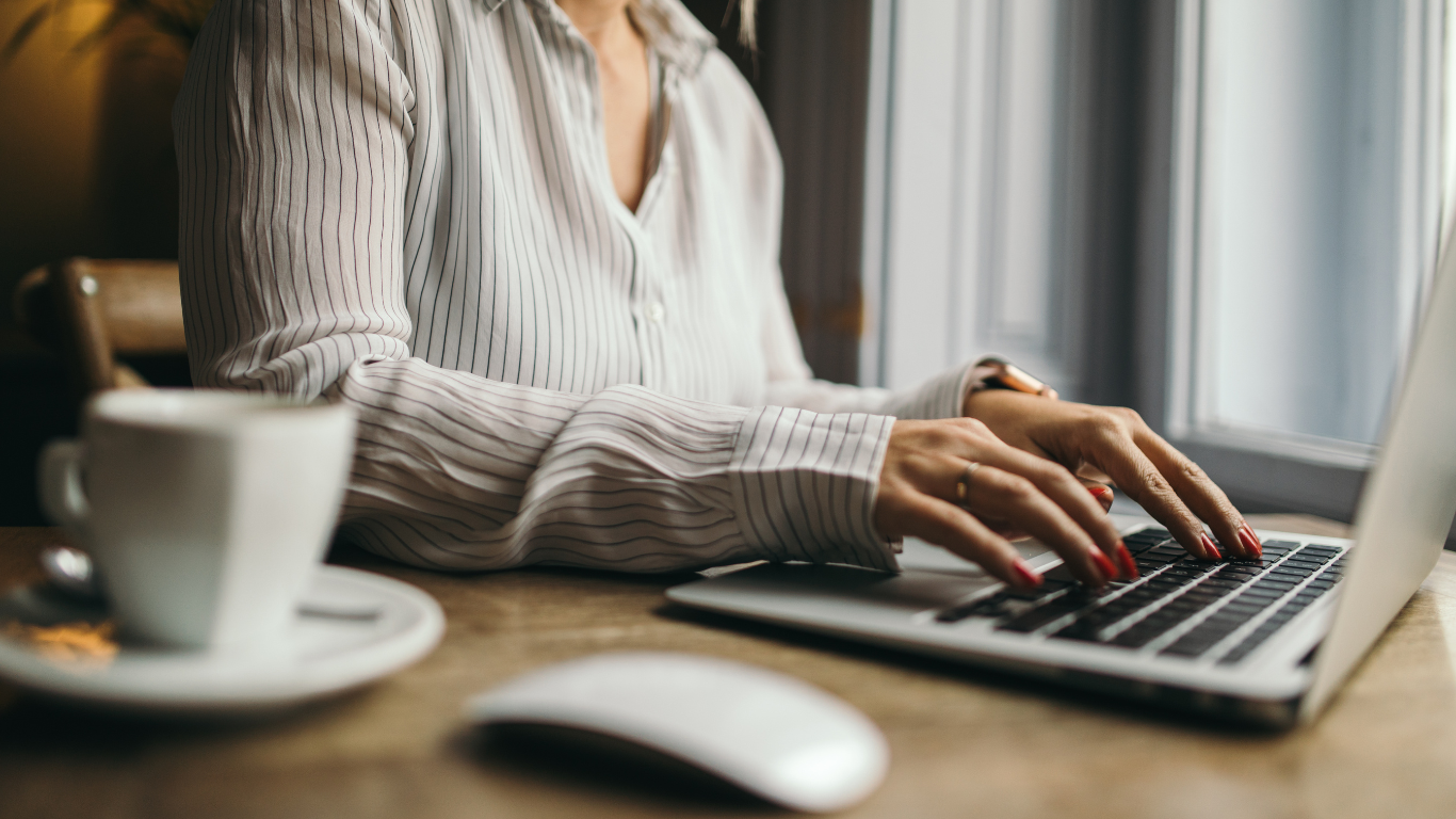 A Virtual Assistant is typing on a laptop computer while sitting at a table with a cup of coffee.
