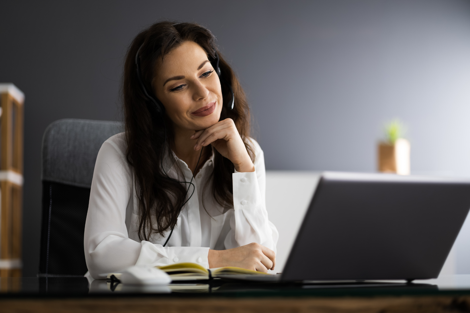A Virtual Assistant wearing headphones is sitting at a desk in front of a laptop computer.