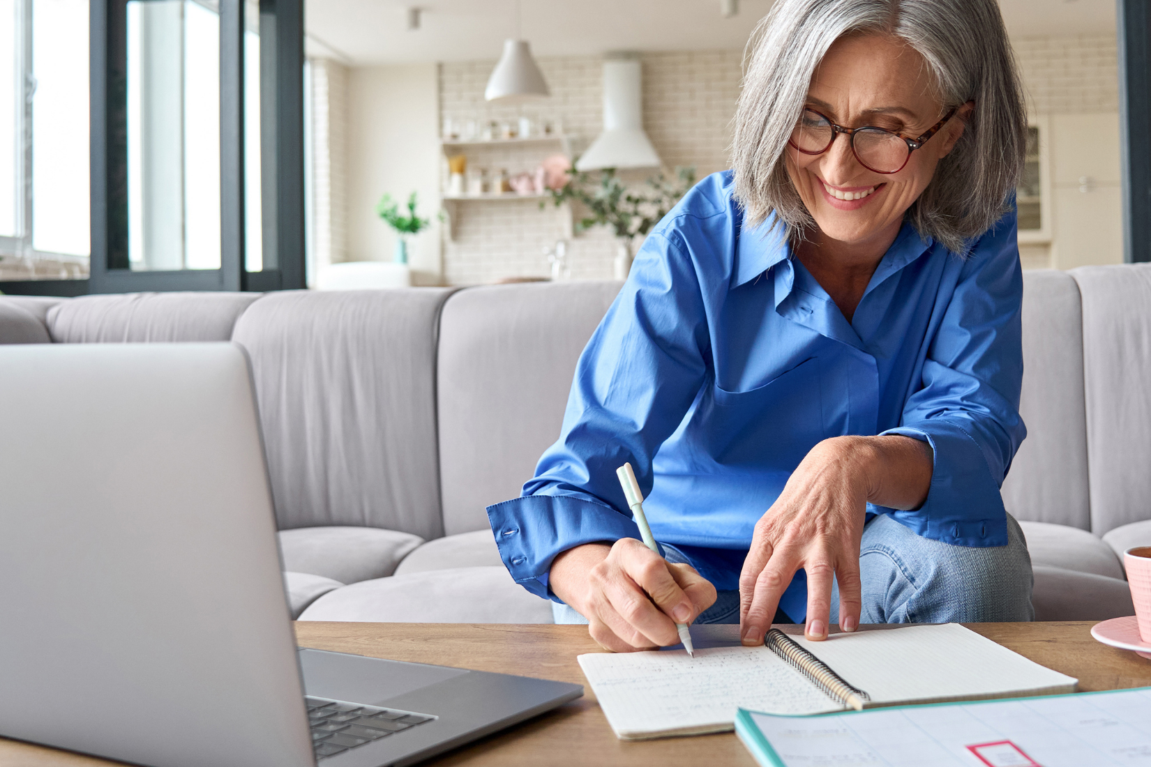 An Virtual Assistant is sitting at a table with a laptop and writing in a notebook.