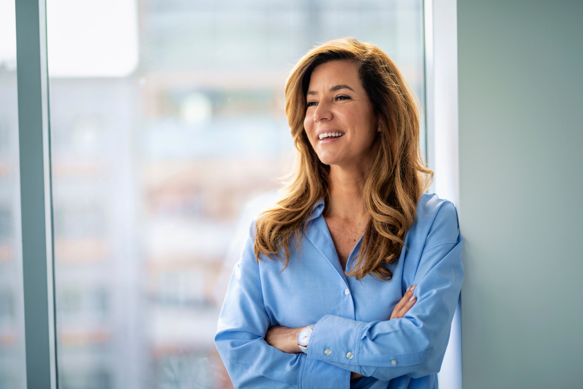 A smiling person with long wavy hair, wearing a light blue button-up shirt, stands with arms crossed by a window.