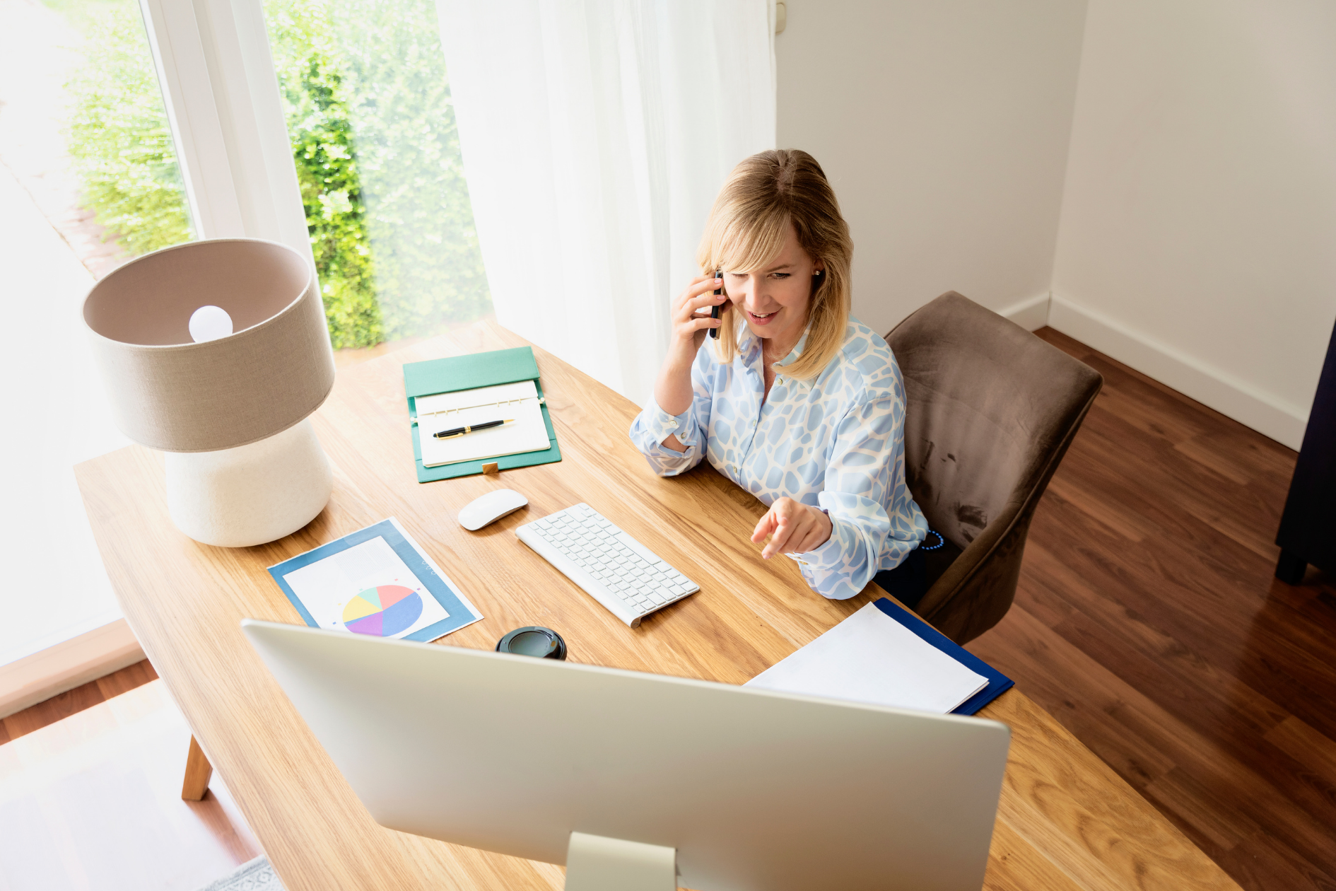A person sitting at a wooden desk in a bright office, talking on a phone while looking at a computer monitor.