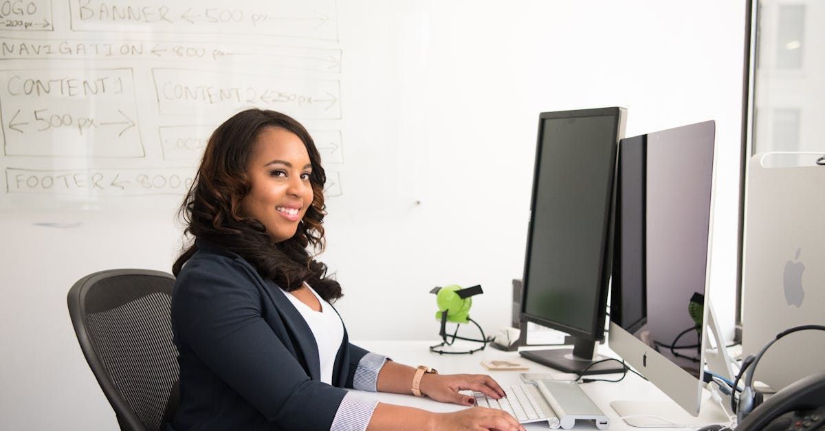 Woman sitting at a desk, smiling while using a computer with two monitors in a bright office.