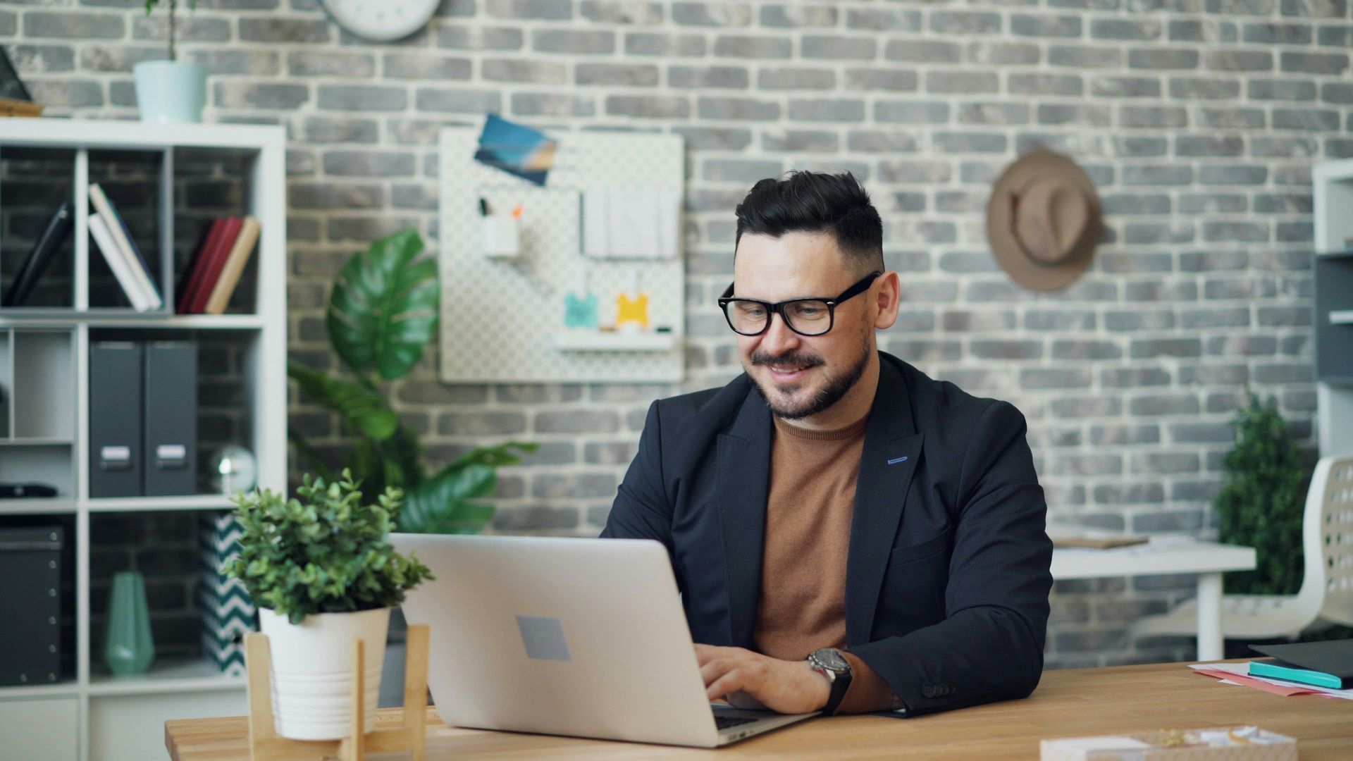 Man in glasses, blazer at desk using laptop, smiling. Brick wall background, office setting.
