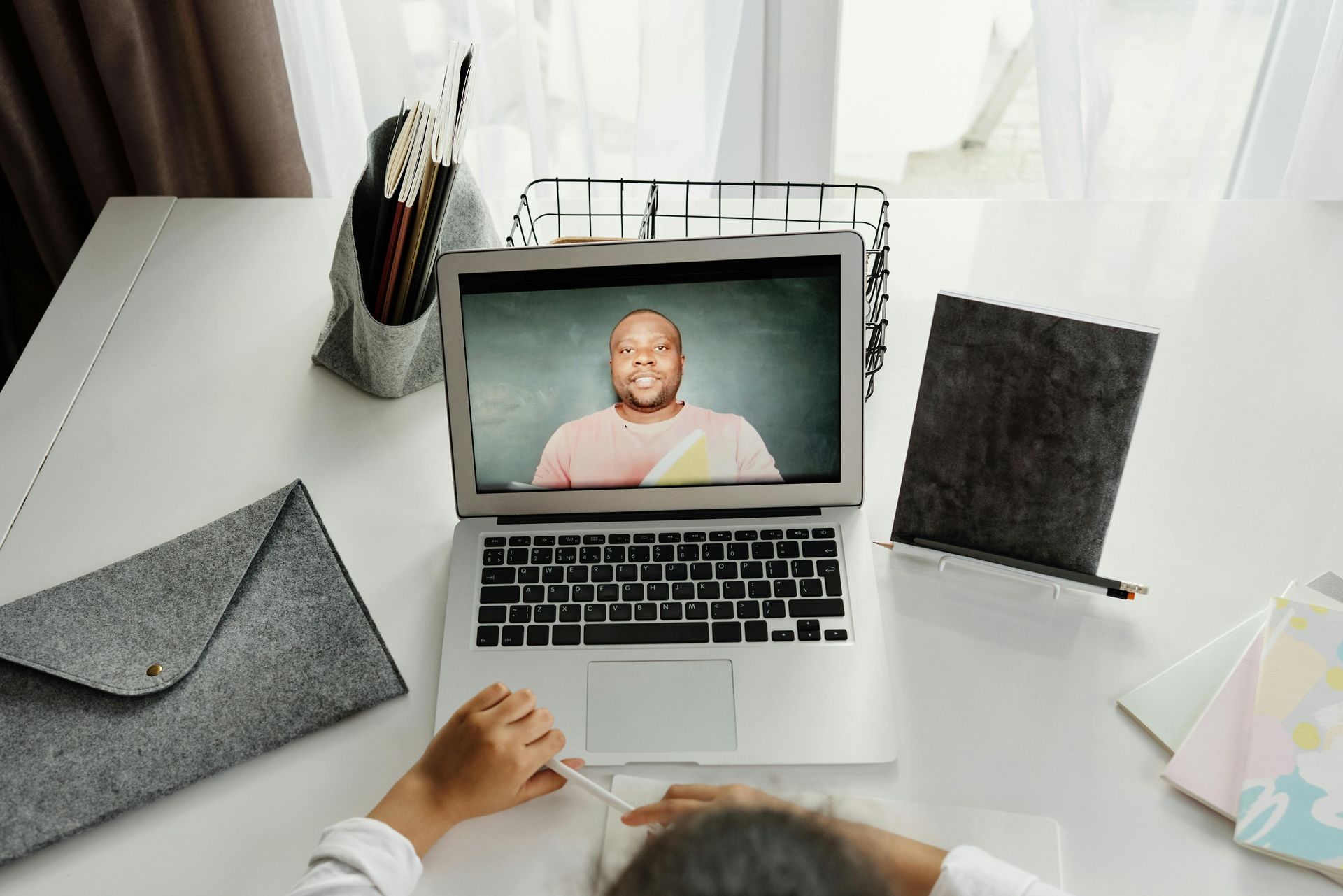 Person at laptop in online meeting, smiling man on screen, white desk, notebooks, pencil case.