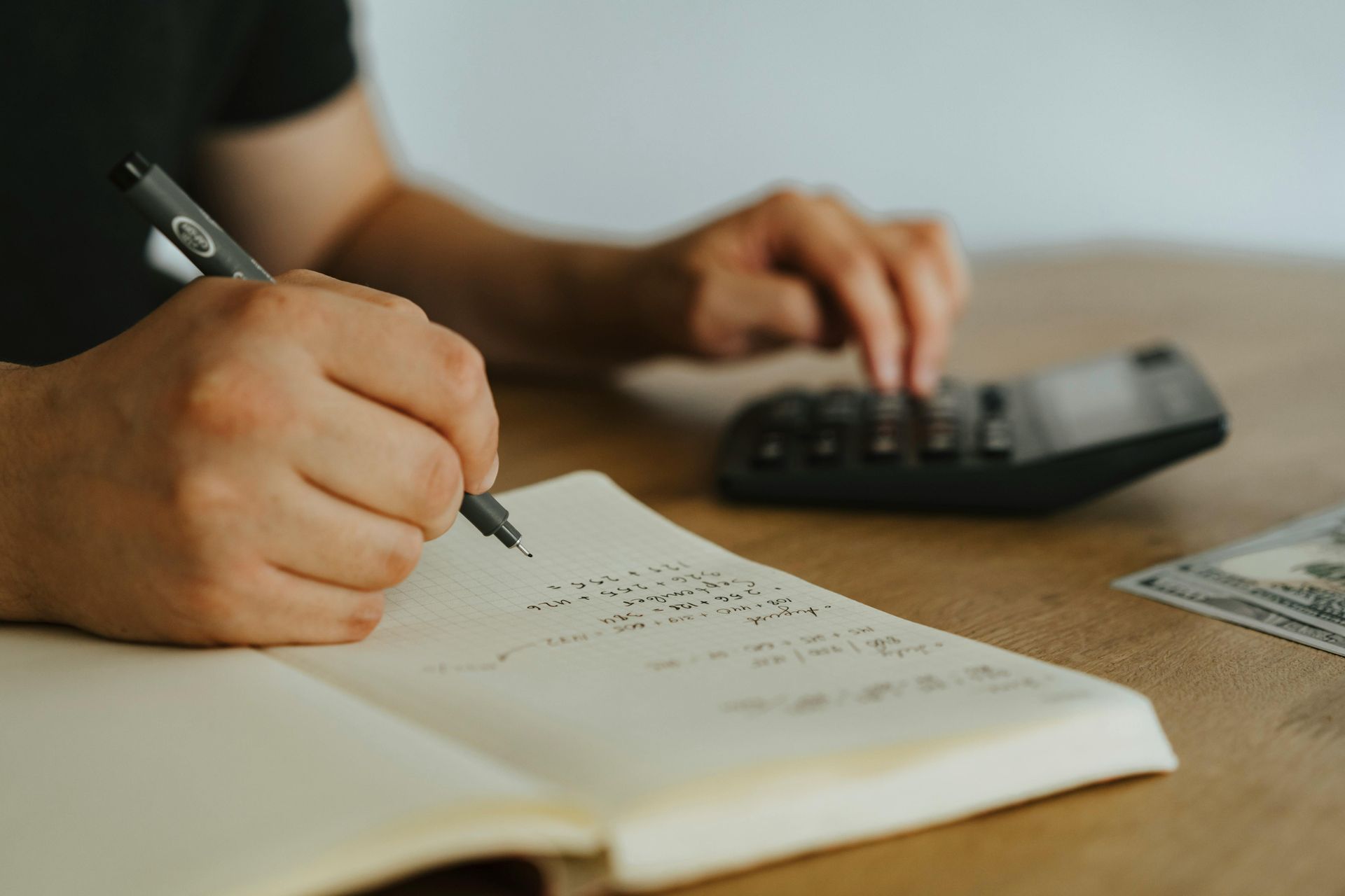 Person using a calculator and writing in a notebook, next to a stack of cash on a wooden table.
