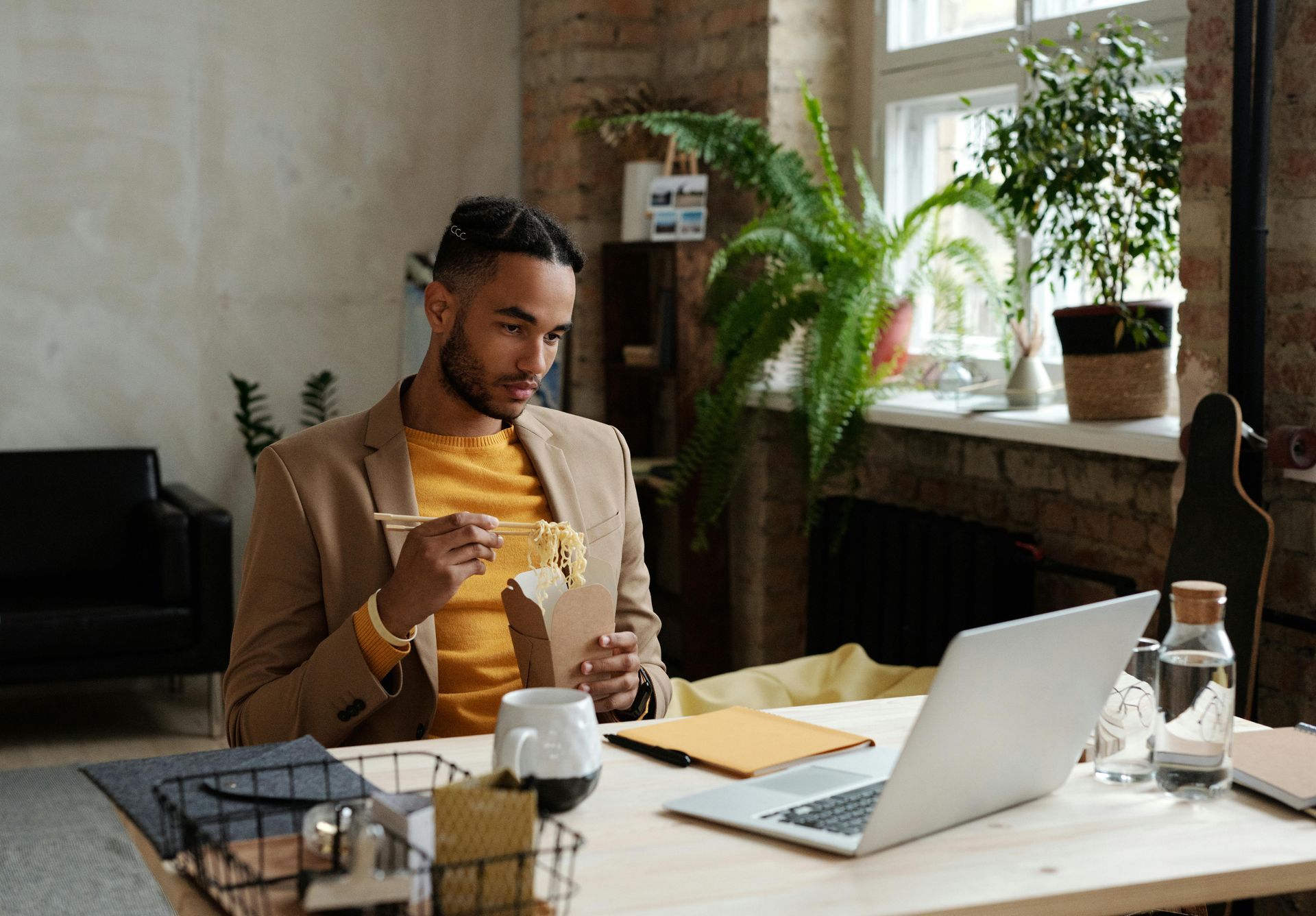 Man in blazer eating noodles at a desk, using a laptop and looking focused. Indoor setting, plants in background.