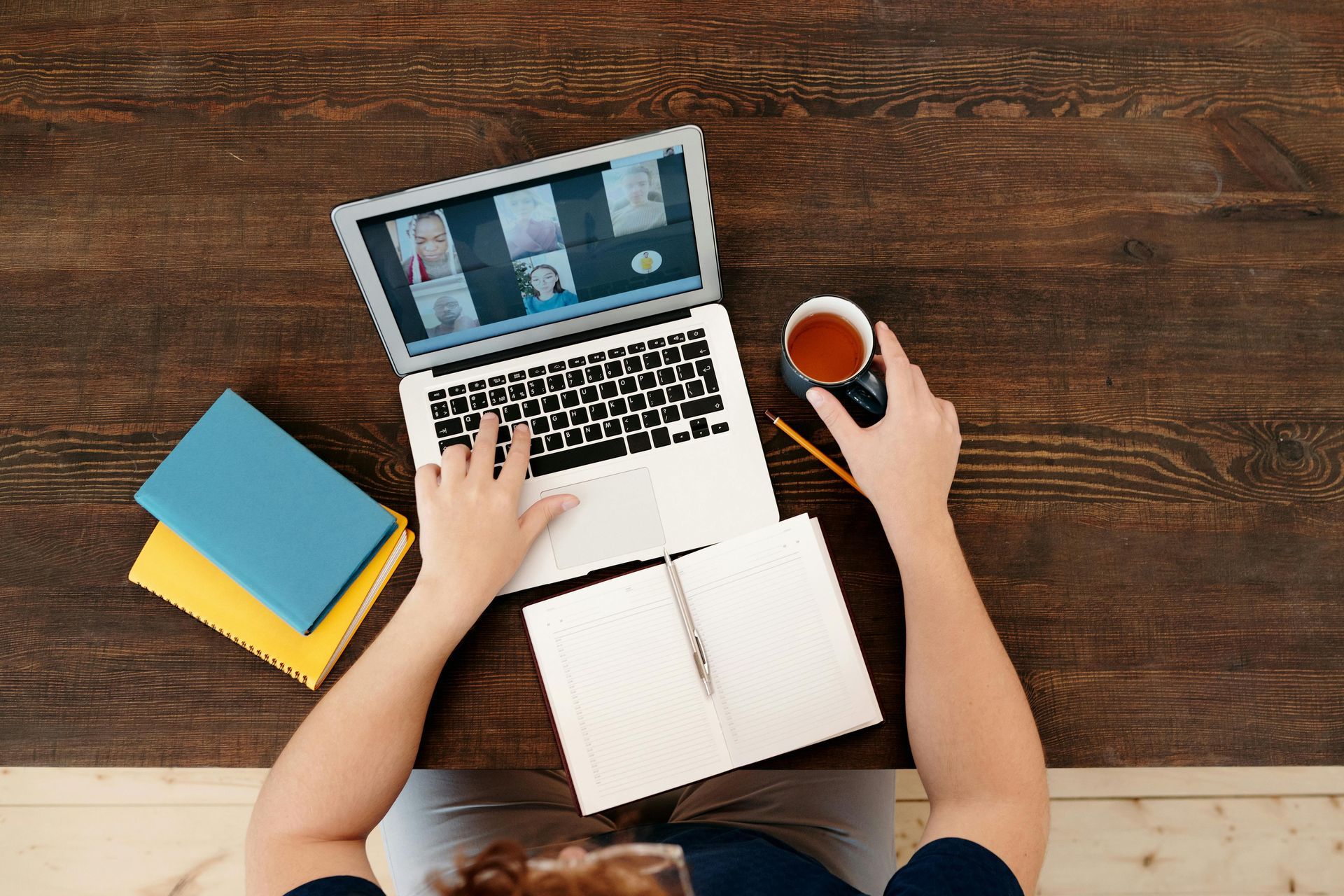 Person using laptop for video call on wooden desk, holding mug, with notepad and books.