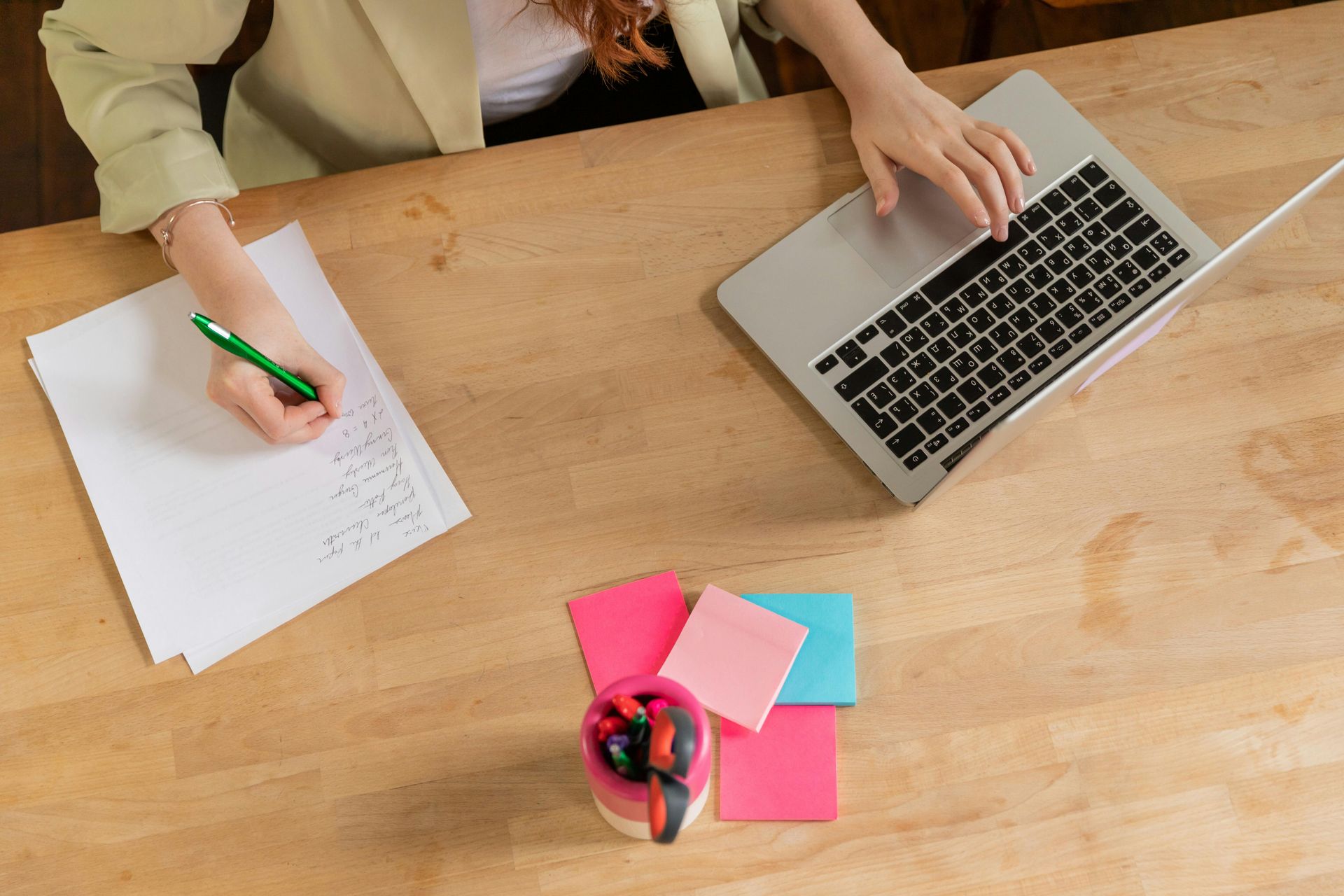 A woman is sitting at a desk using a laptop computer and writing on a piece of paper.