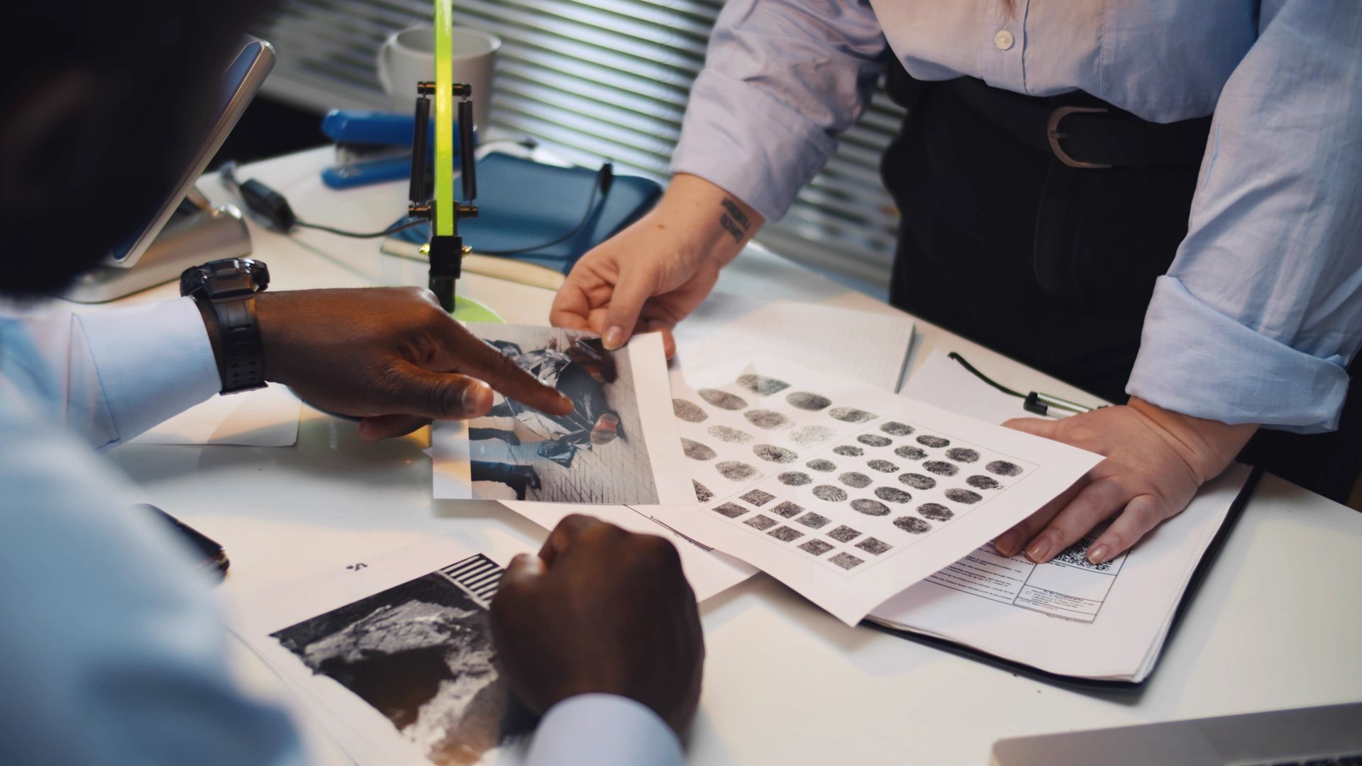 Two people examining photos and a printout at a desk. One points at a photo; the other holds documents.