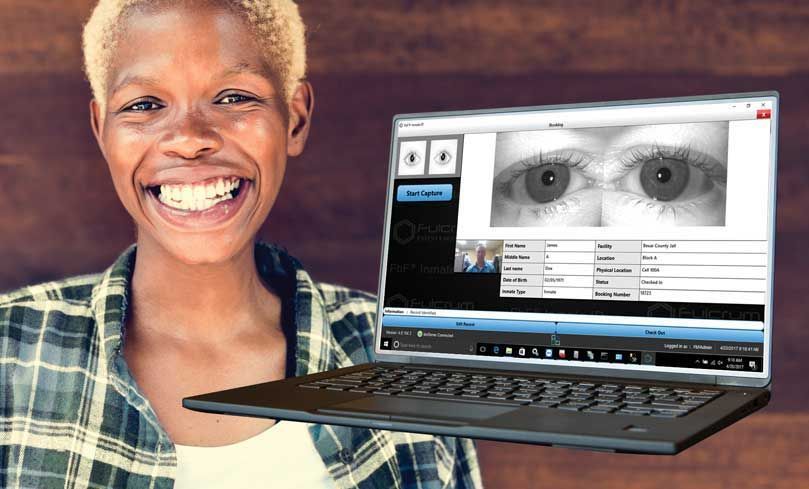Woman smiling, holding a laptop displaying an eye exam. Brown background.
