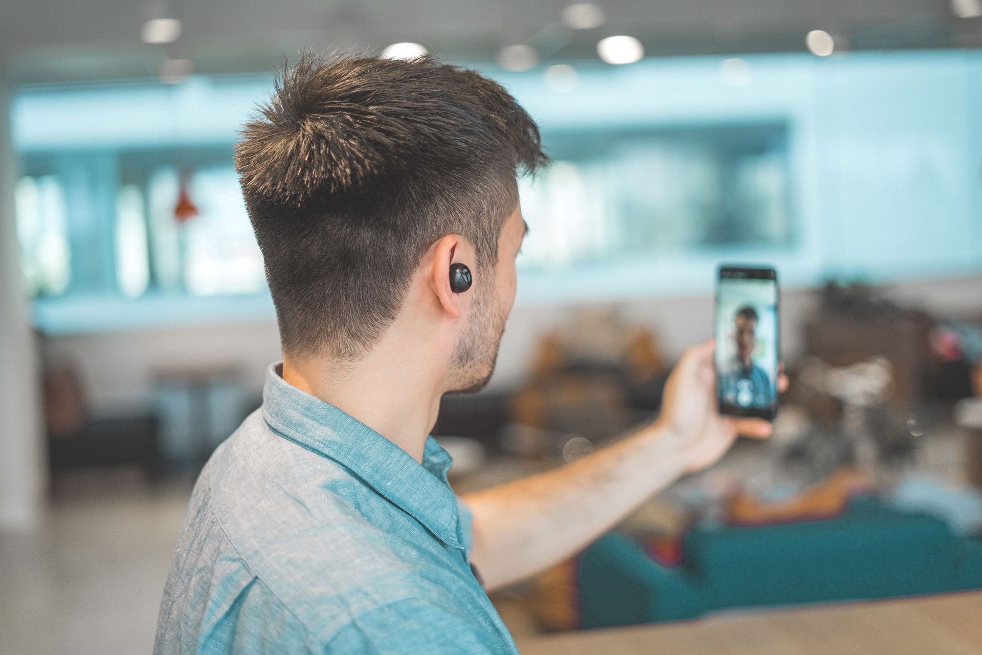 Person wearing earbuds, holding phone, video-calling someone, in a bright office space.