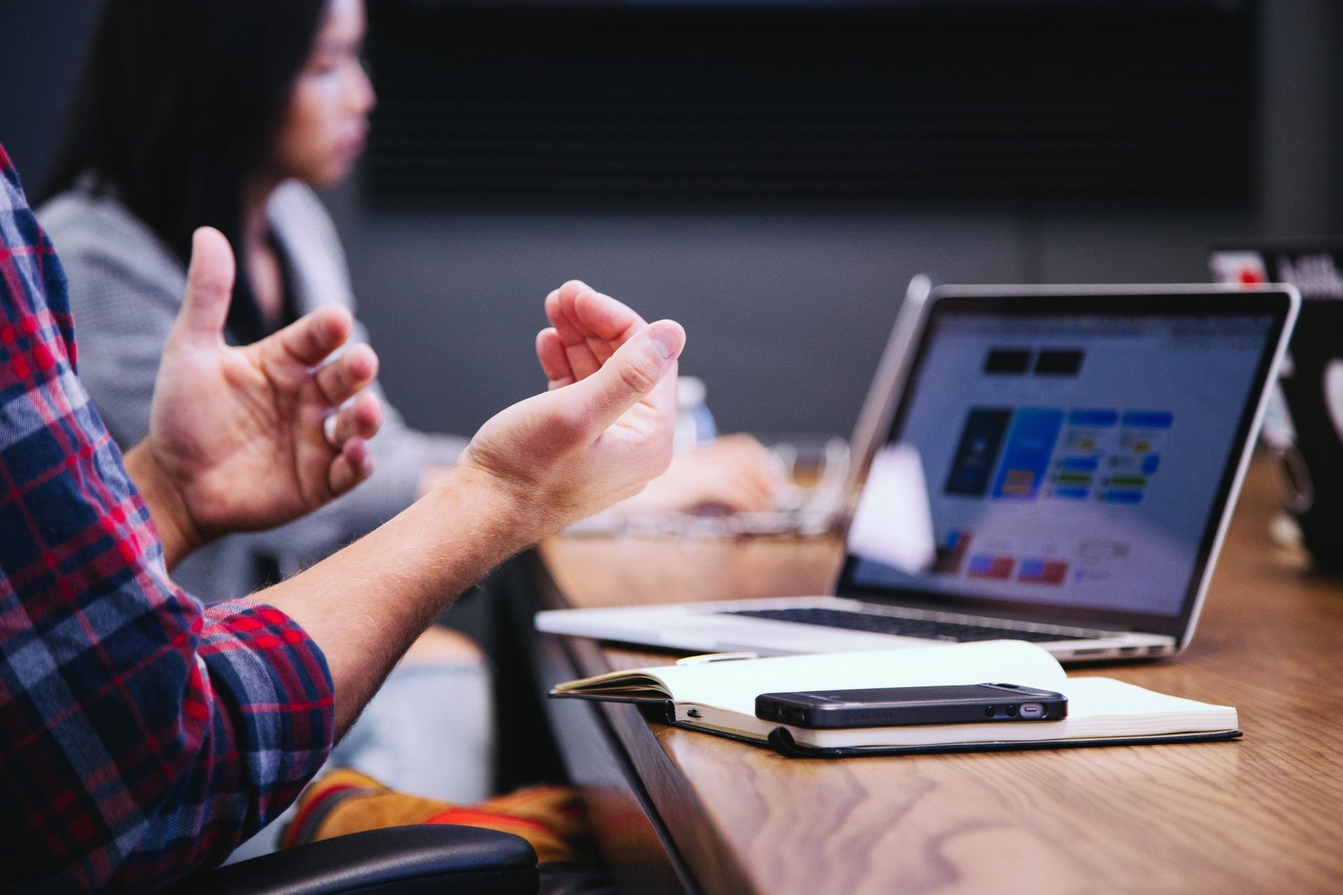 Person gesturing with hands during a meeting, laptop and notepad on table.