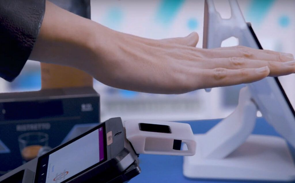 Hand hovering over a white device, possibly for payment, in a store setting. Other tech and screens visible.