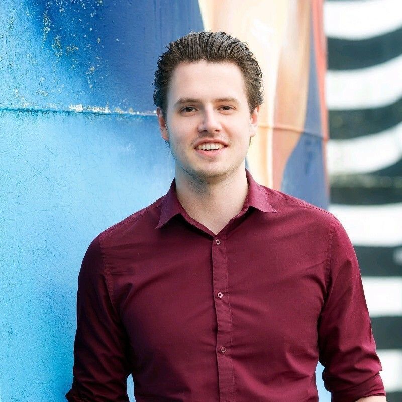 Man in a burgundy shirt smiles, leans against a blue and colorful wall.