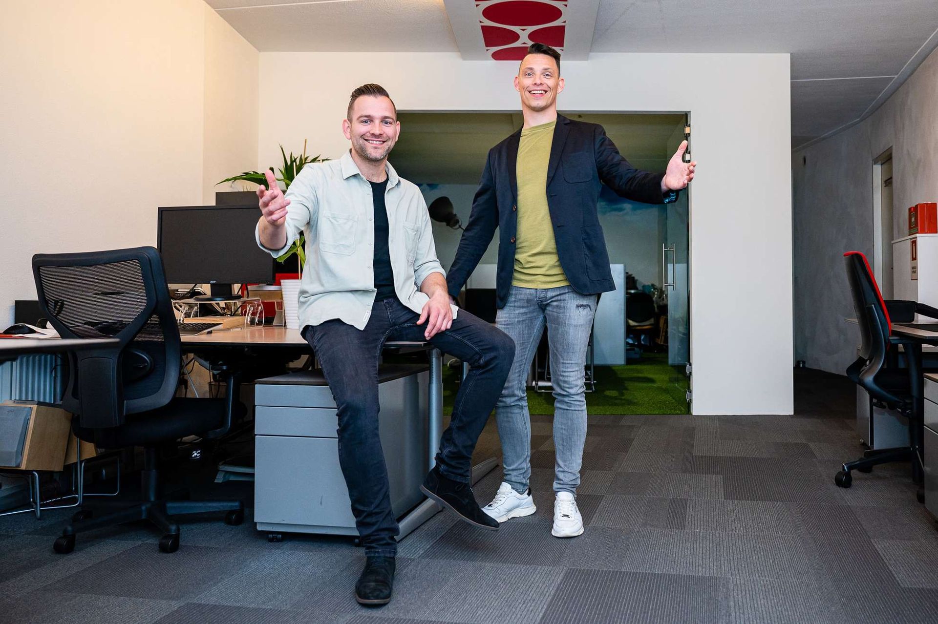 Two men pose in office, one seated on desk, both smiling and gesturing, indoors.