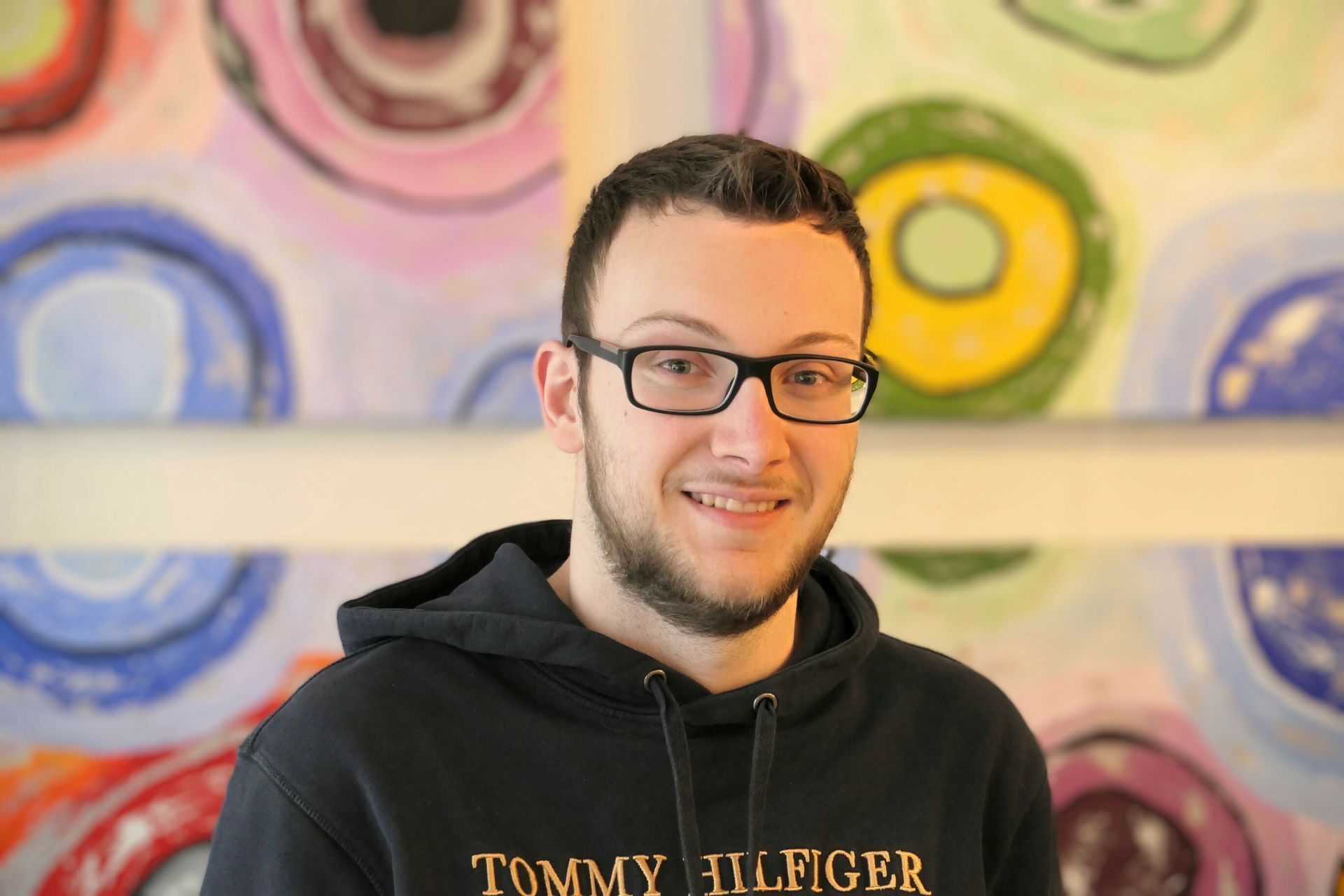 Man with glasses smiles, wearing a black Tommy Hilfiger hoodie, in front of colorful abstract art.
