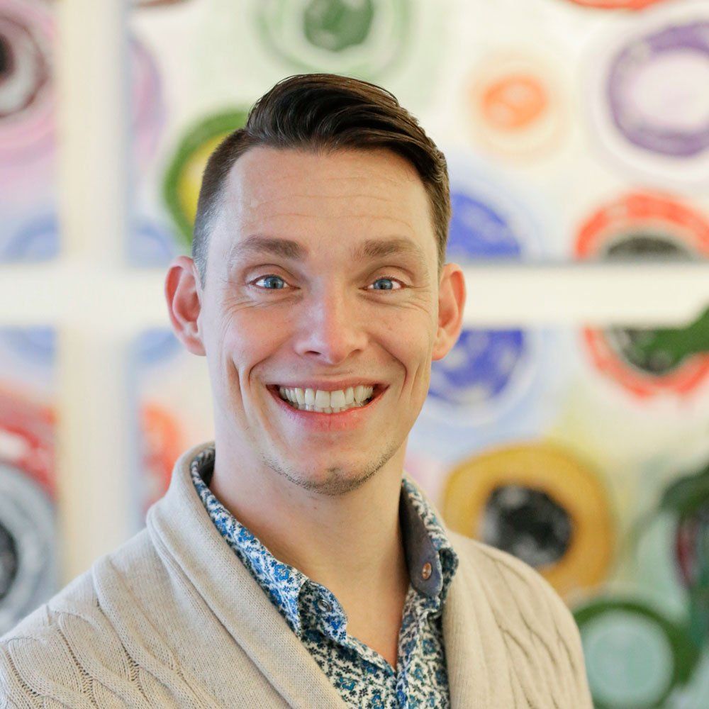 Man with short dark hair, smiling, wearing a light cardigan and patterned shirt, with colorful background.
