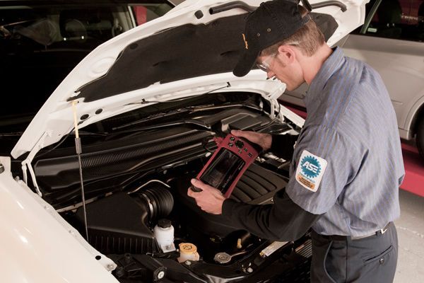 Mechanic in uniform with a black cap using a diagnostic tool on a car engine in a garage.