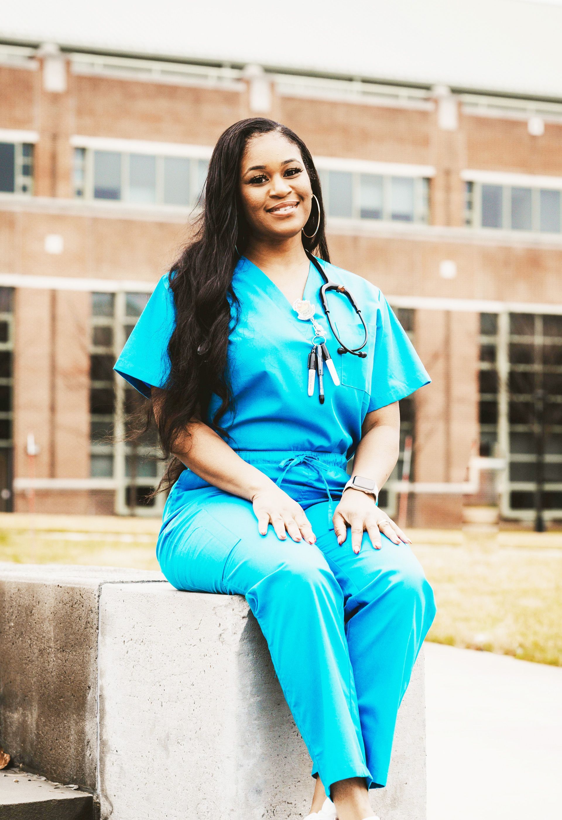 Nurse in teal scrubs sits outside, smiling. Stethoscope around her neck.