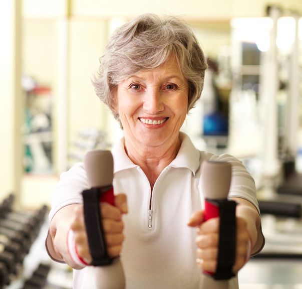Smiling older woman in gym, holding up dumbbells.