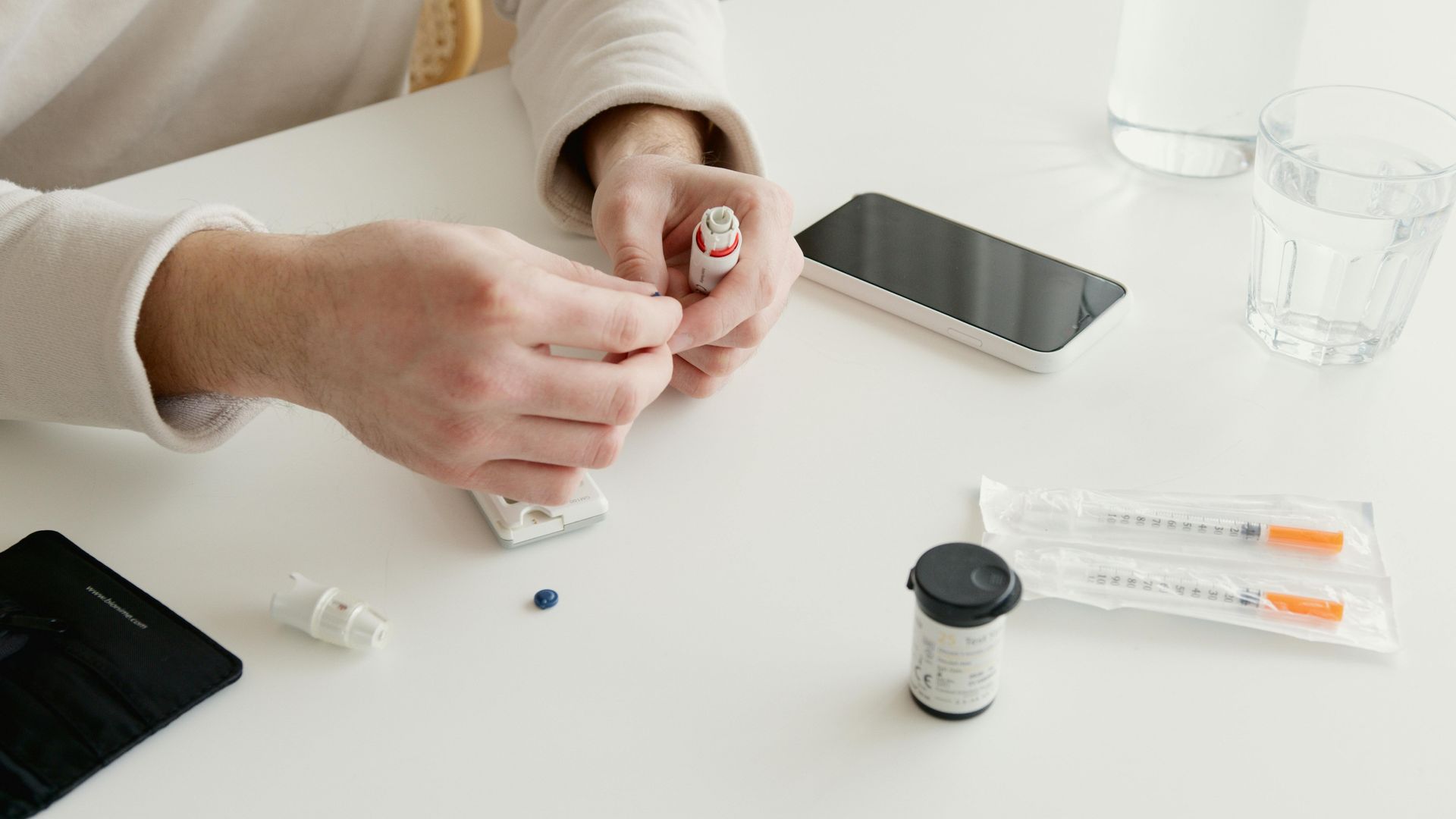 Person preparing a blood glucose test, holding a lancet device and test strips on a white table.