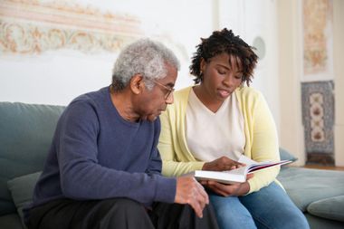 An elderly man and a young woman looking at a book together on a sofa.