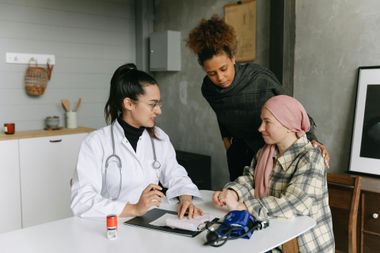 A doctor consulting with a patient wearing a headscarf, accompanied by a woman, in an office.