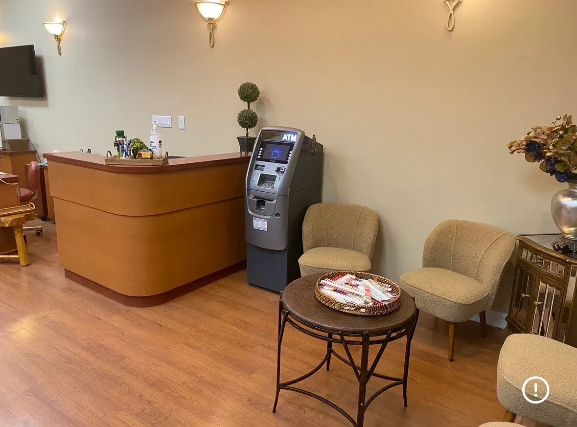 Salon with front desk, ATM, two beige chairs, and a small round table with flowers and magazines.