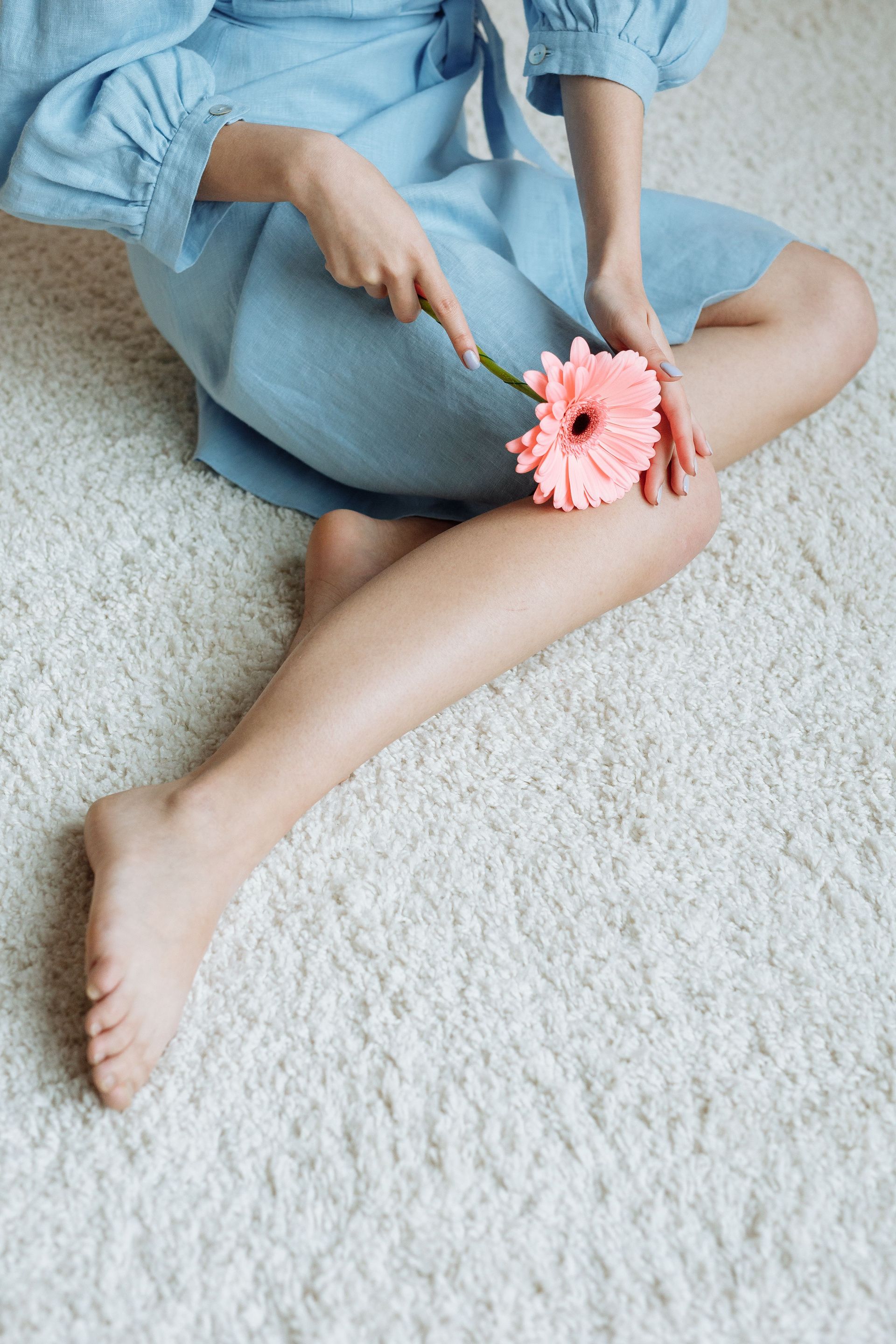 Child in a blue dress sitting on a white rug, holding a pink flower against their leg.