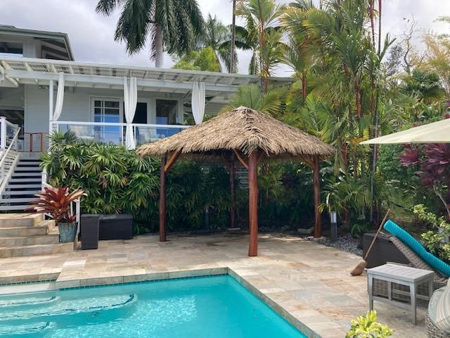 A thatched gazebo sits next to a swimming pool in front of a house.