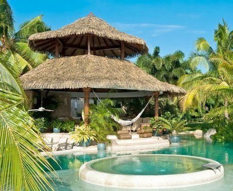 A hut with a thatched roof sits next to a swimming pool surrounded by palm trees.