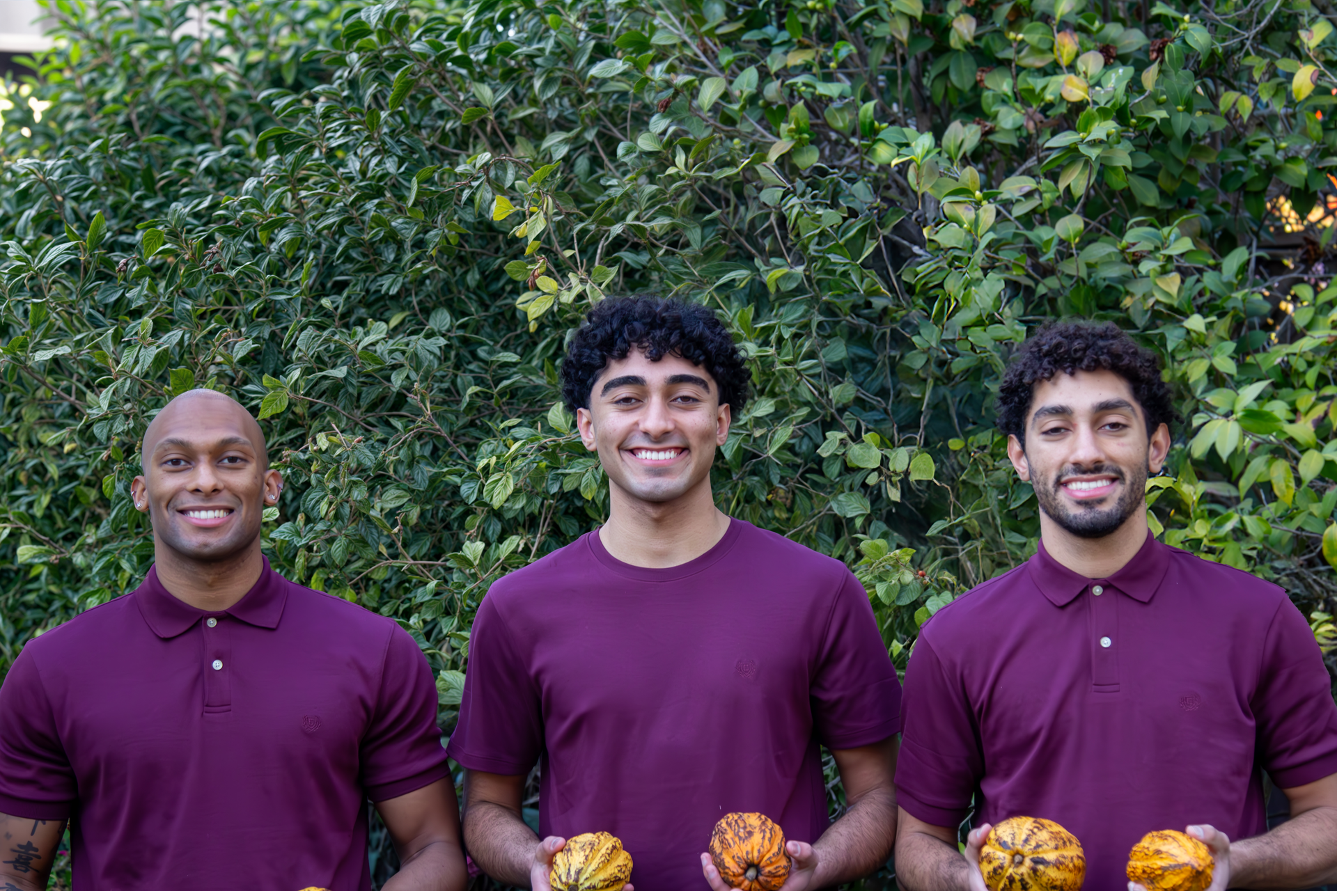 Three men wearing purple polo shirts are holding a few fruits in their hands
