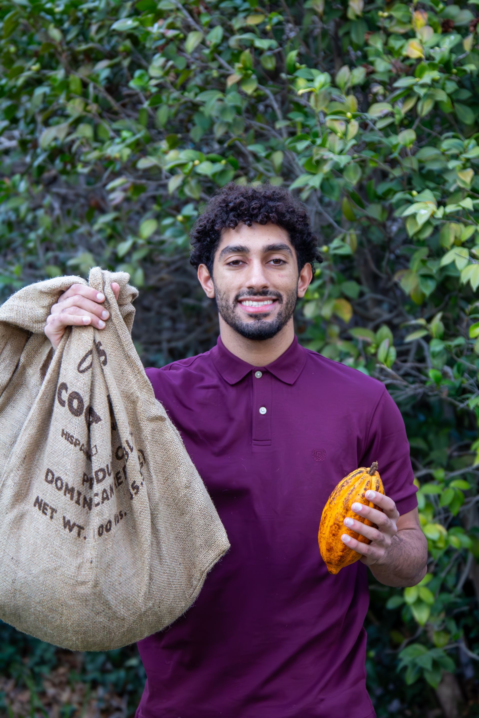 A man in a purple shirt is holding a bag of cocoa beans.