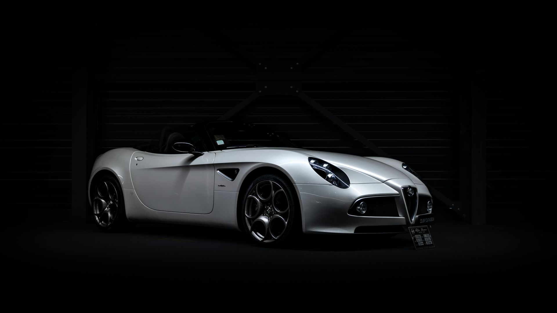 White Alfa Romeo 8C Spider convertible car, dark setting, headlights illuminated.