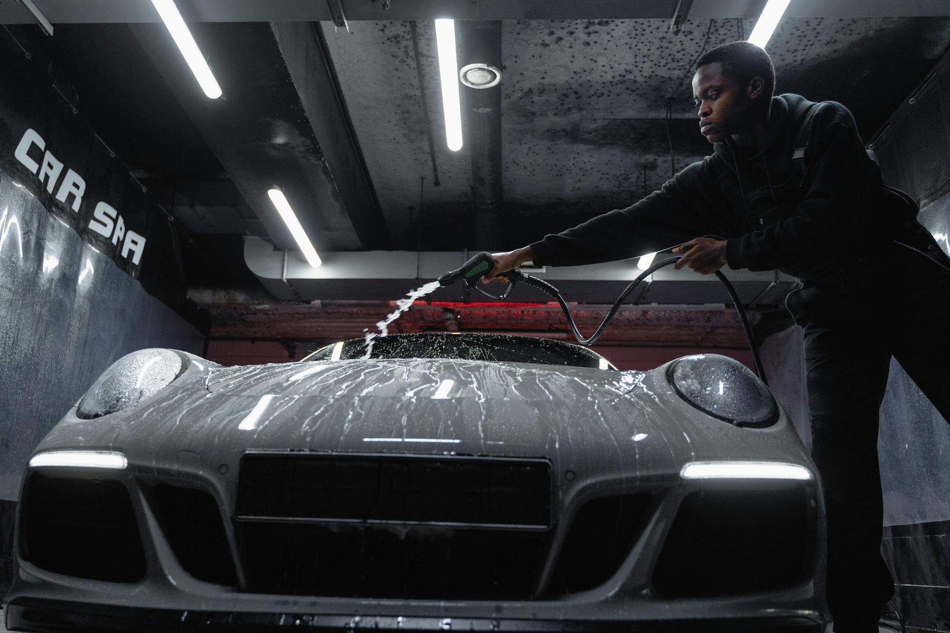 Man washing a silver sports car in a dimly lit garage, spraying water with a hose.