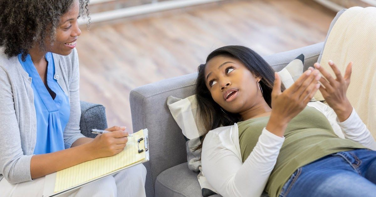A person lying on a couch gestures while speaking to a professional holding a clipboard and pen in a seated session.