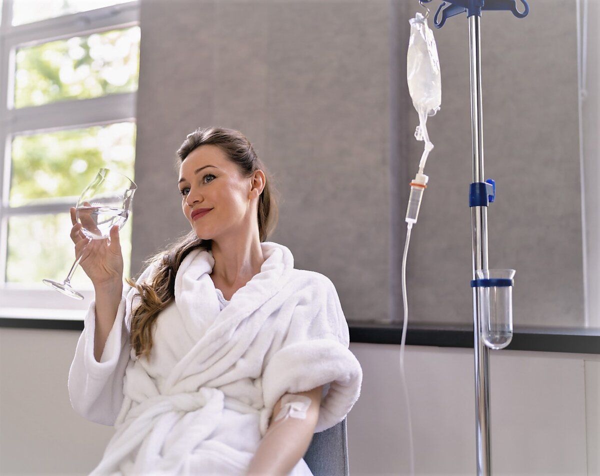A person in a white bathrobe receives an IV drip while holding a wine glass in a bright room.