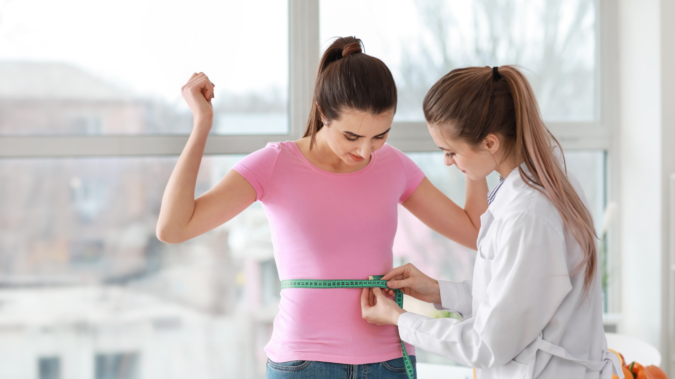 A person in a lab coat measures another person's waist with a green tape measure in a bright office.