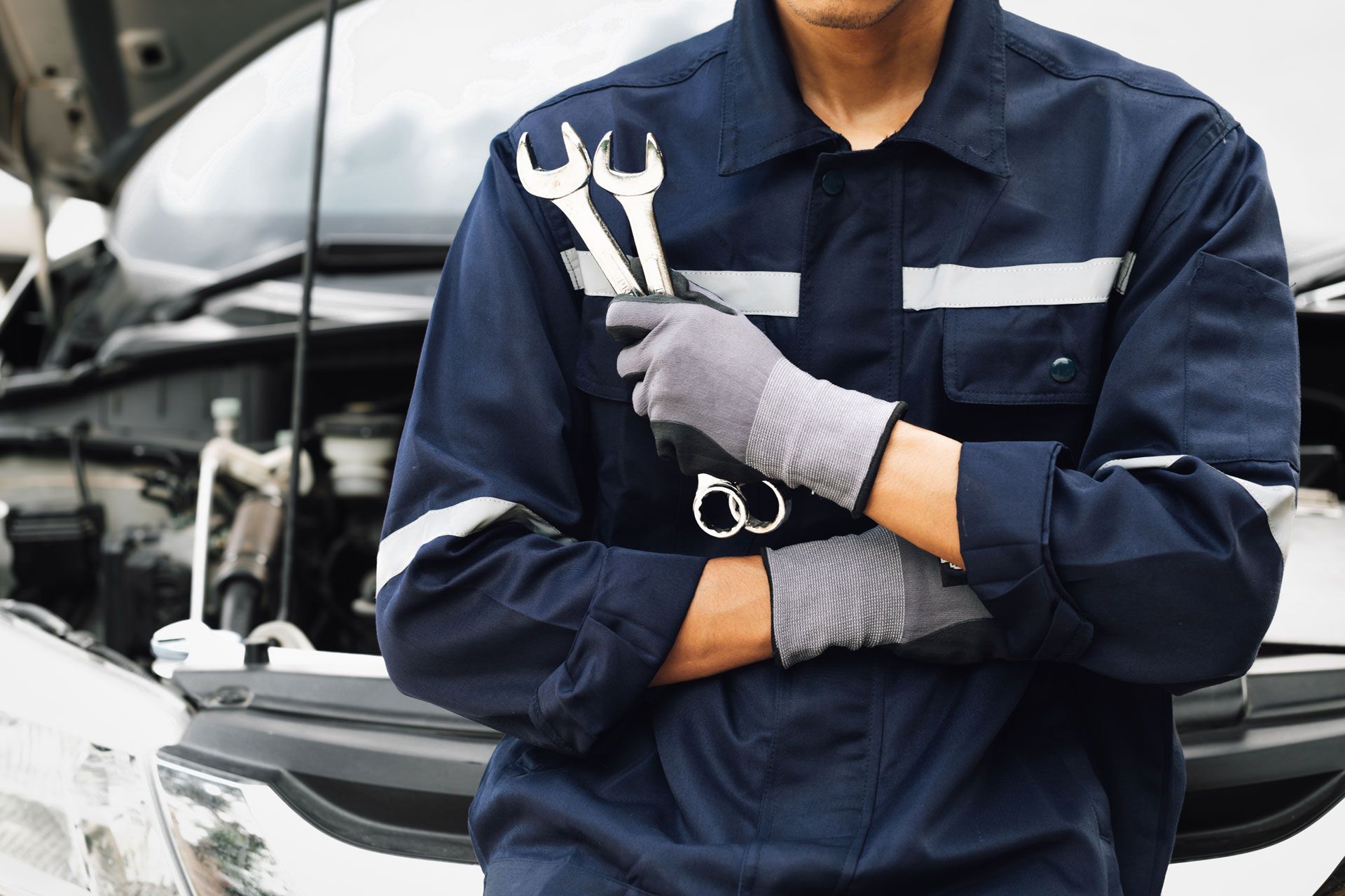 Mechanic in navy coveralls holding wrenches in front of a car with its hood open