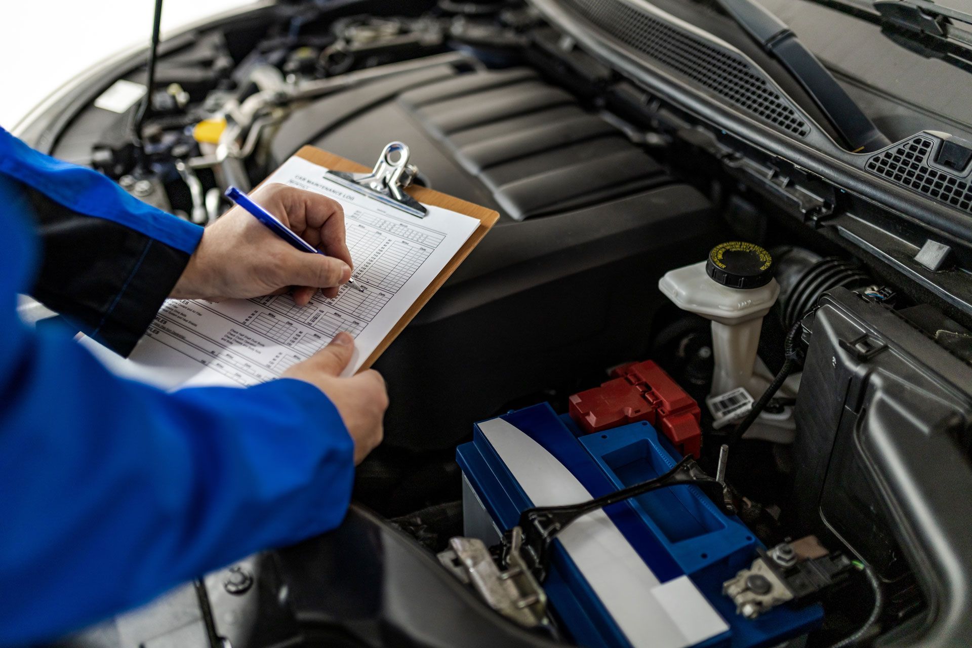 Mechanic writing on a clipboard beside an open car hood with exposed engine and battery