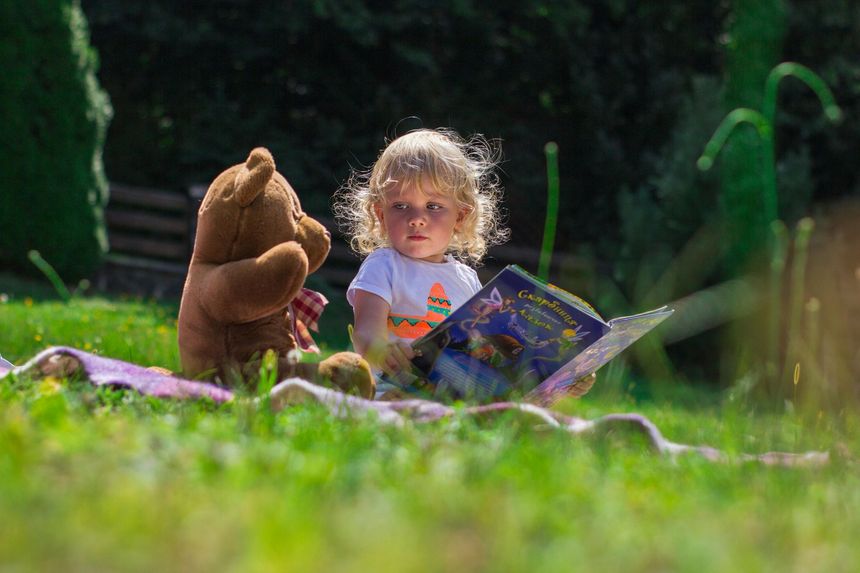 A young child sitting on the grass, reading a book to her teddy bear