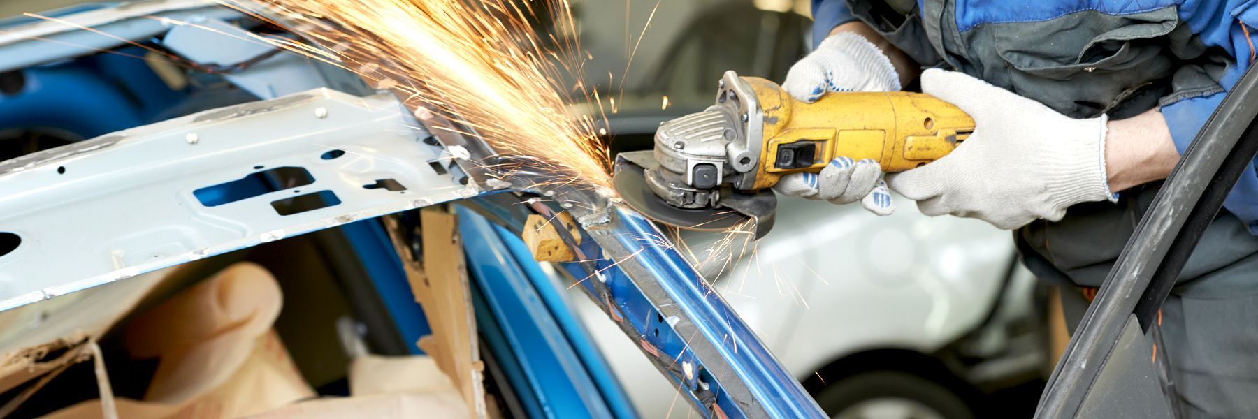 A person using a yellow grinder on car metal, creating sparks. They wear gloves and a blue uniform.