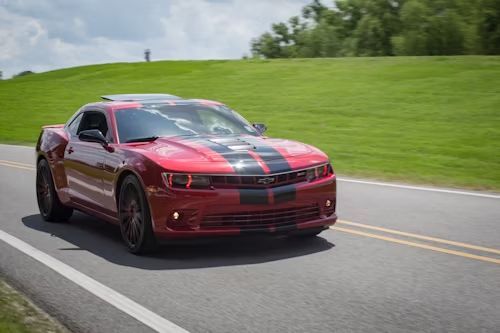 Red Chevrolet Camaro with black racing stripes on a paved road, green hill in the background.