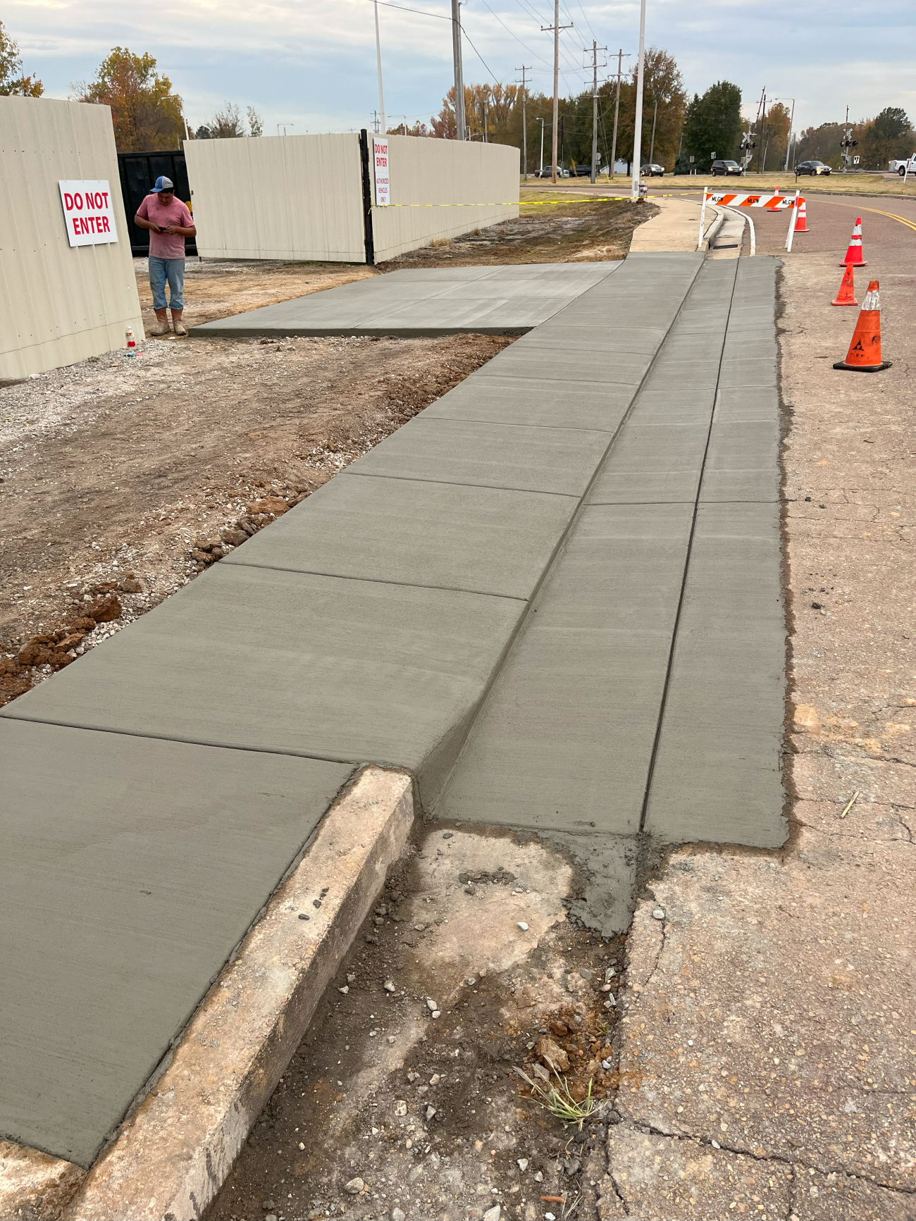 New concrete sidewalk being constructed near a fence and road with a worker in the background.