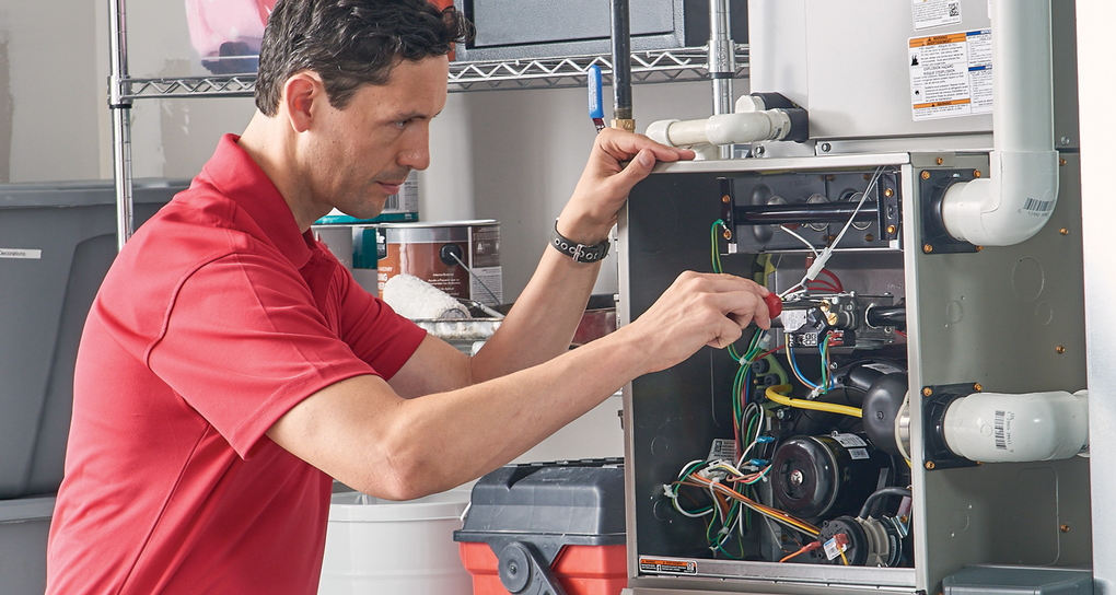 Man in red shirt repairs furnace in a utility room, working on wires and components.