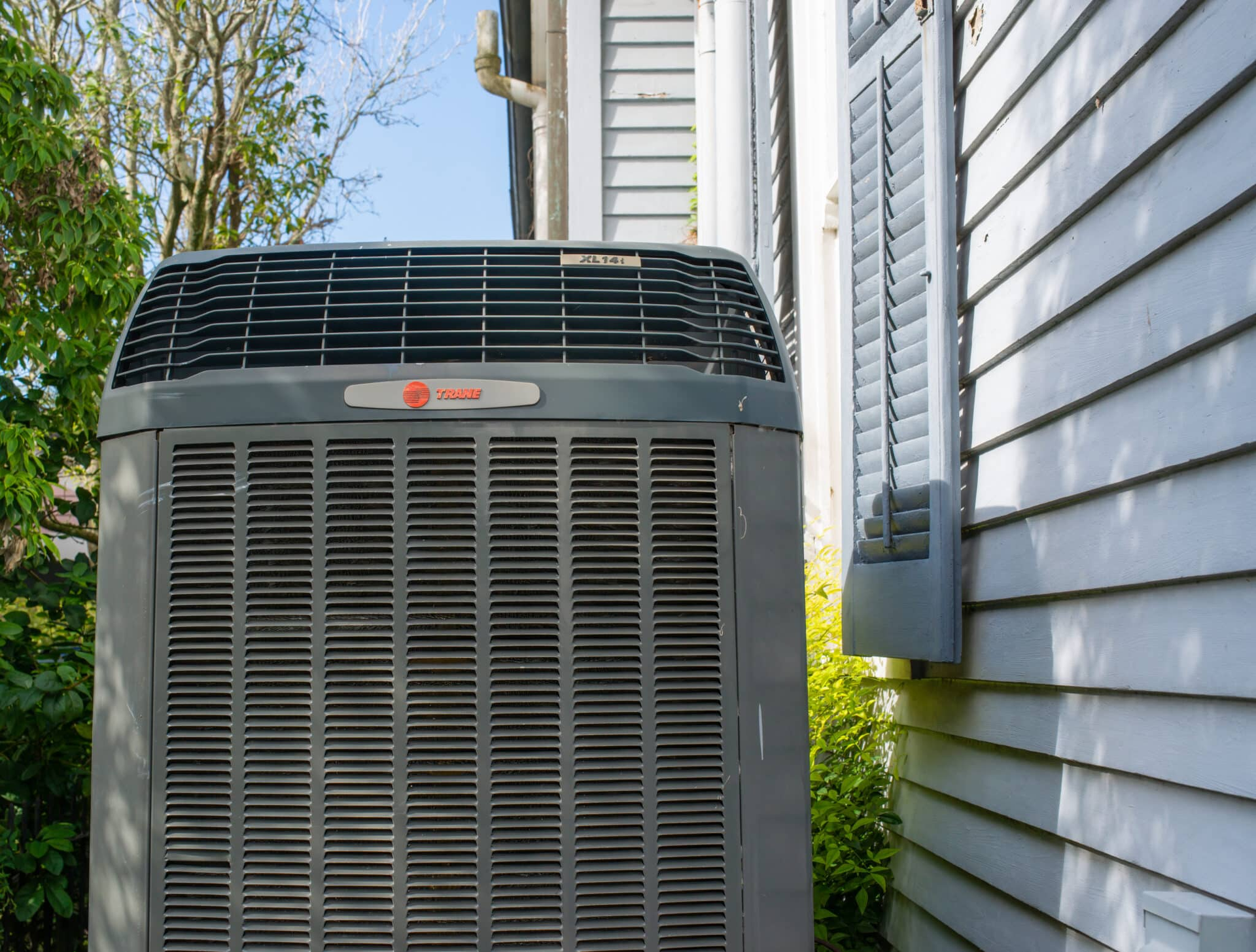 Gray air conditioning unit next to a white house with a shutter; outdoors.