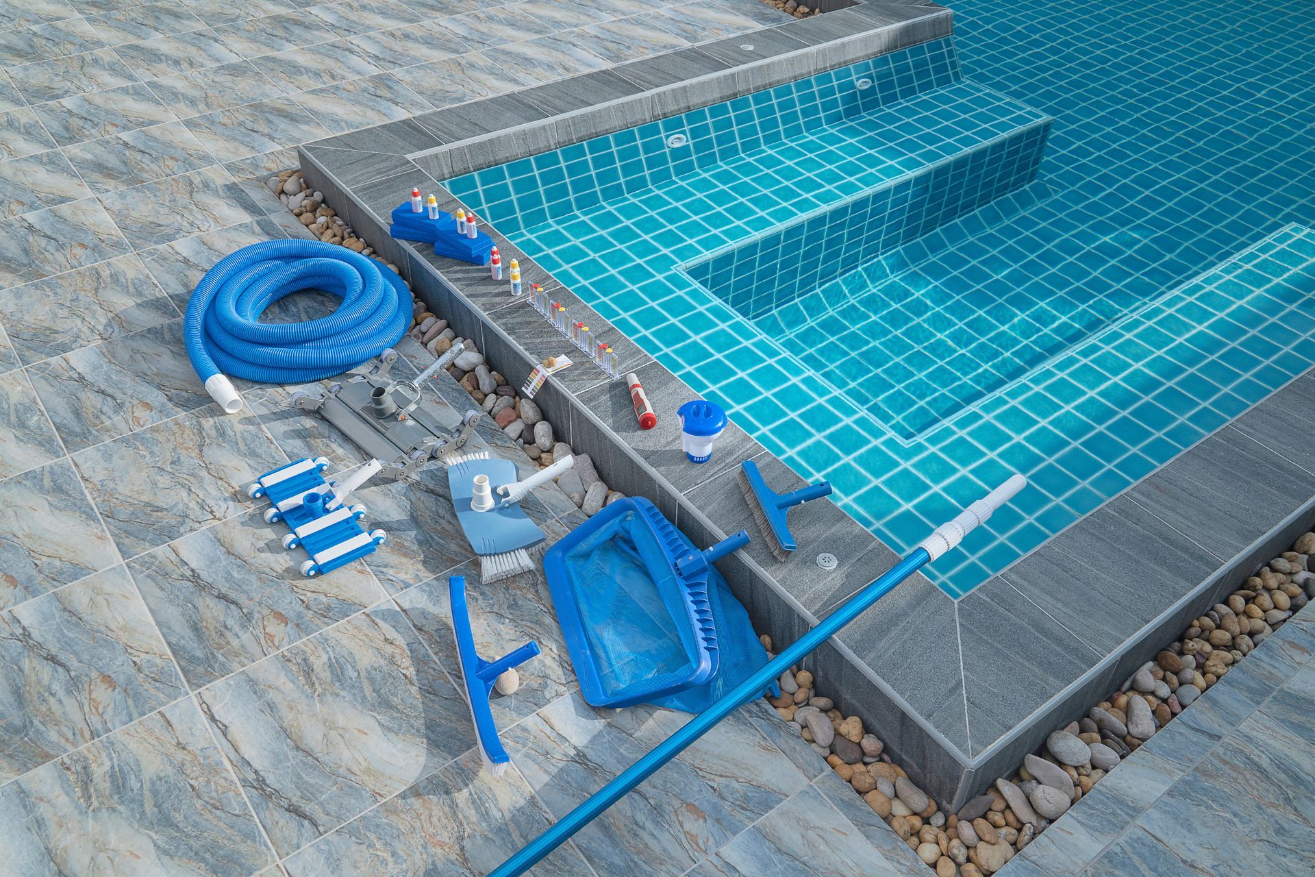 Pool cleaning equipment next to a tiled pool, including vacuum, skimmer, brush, and hose.