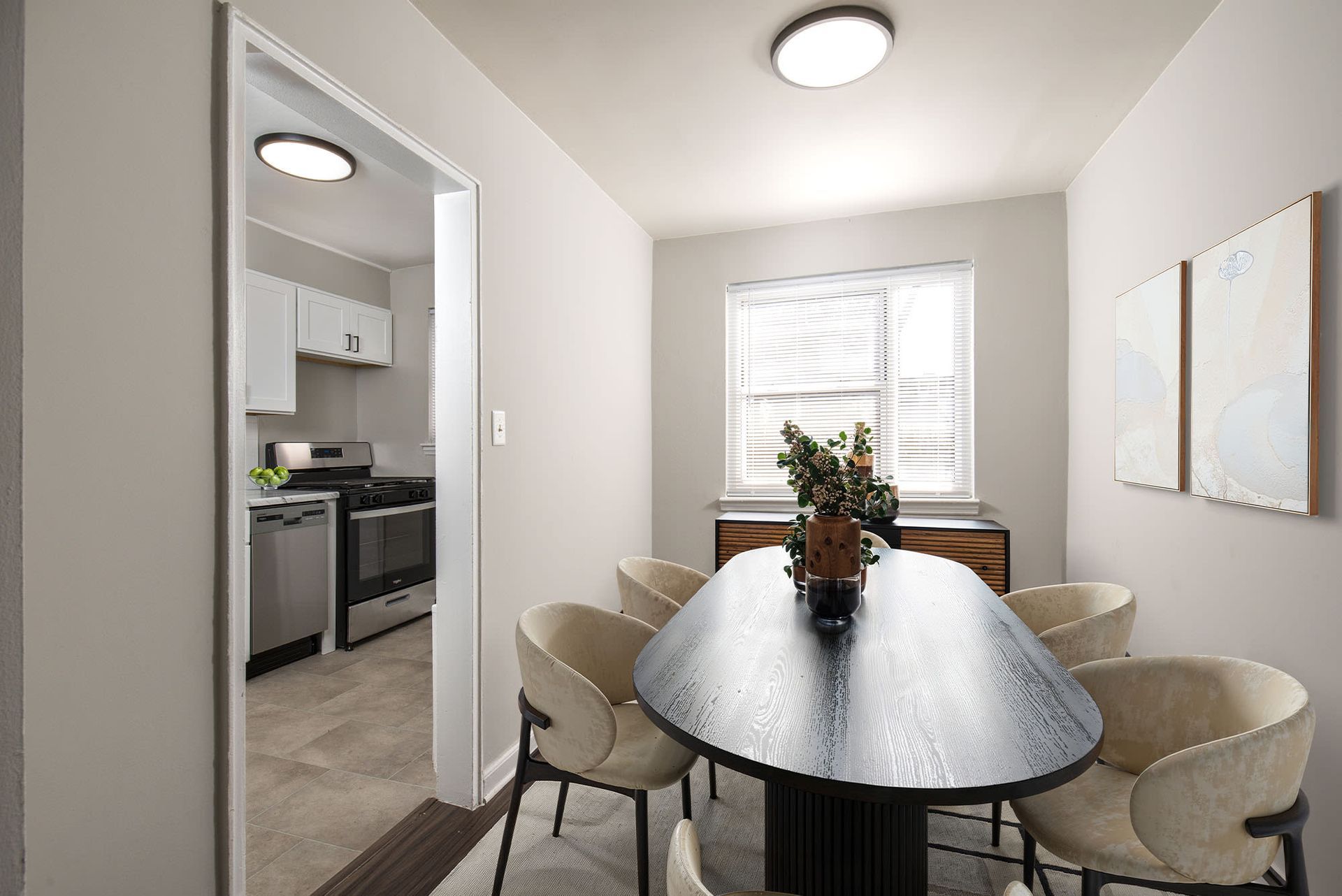Dining area with an oval wooden table and six cream chairs, adjacent to a small kitchen.